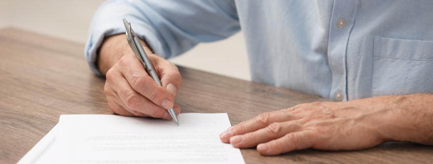 Senior man signing Last Will and Testament at wooden table, closeup