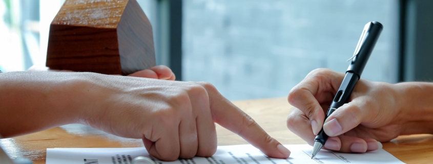 Hands signing a property agreement beside a set of keys and a wooden model house